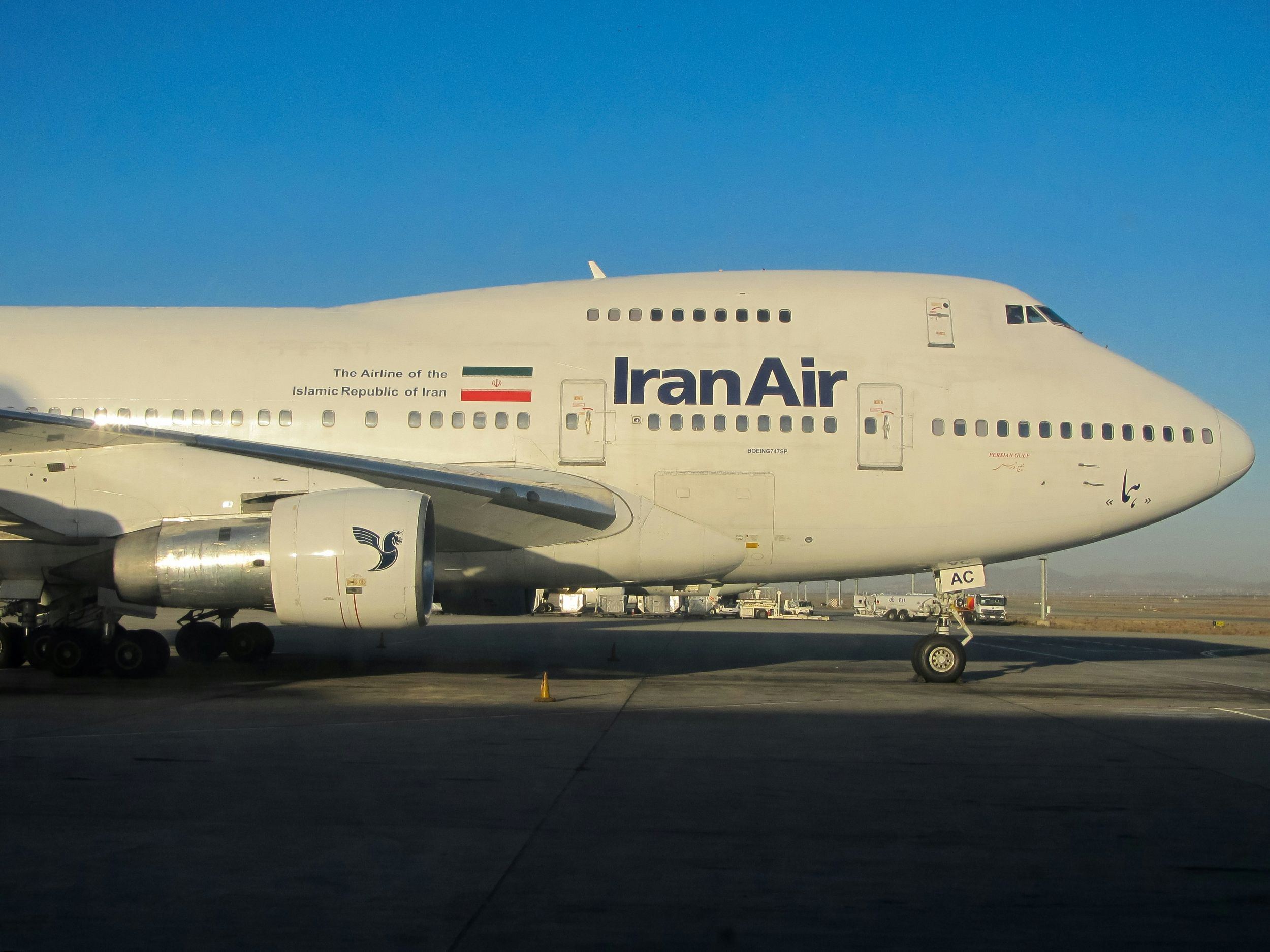The front section side profile of a Boeing 747. The nose faces the right and it in the far centre right of the image while the centre left of the image pictures the body and wing extending beyond the camera's scope. The background is bright blue sky with no clouds, and the foreground depicts the grey concrete of a runway, shadowed by the large aircraft. The navy blue branding 'IranAir' adorns the side of the aircraft as well as the Iranian flag.