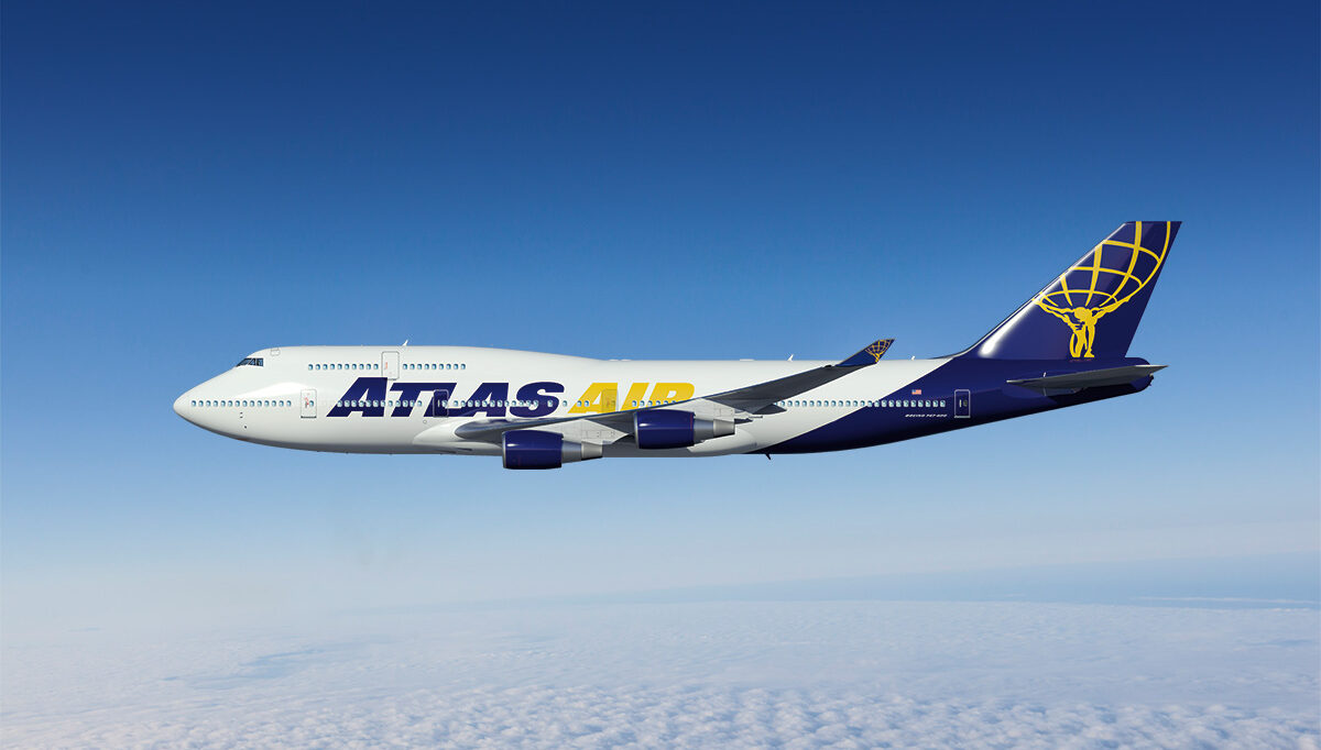 An Atlas Air aircraft flying above the clouds with a blue sky in the background. White and blue livery, with a yellow symbol on the tail of a man holding up earth.