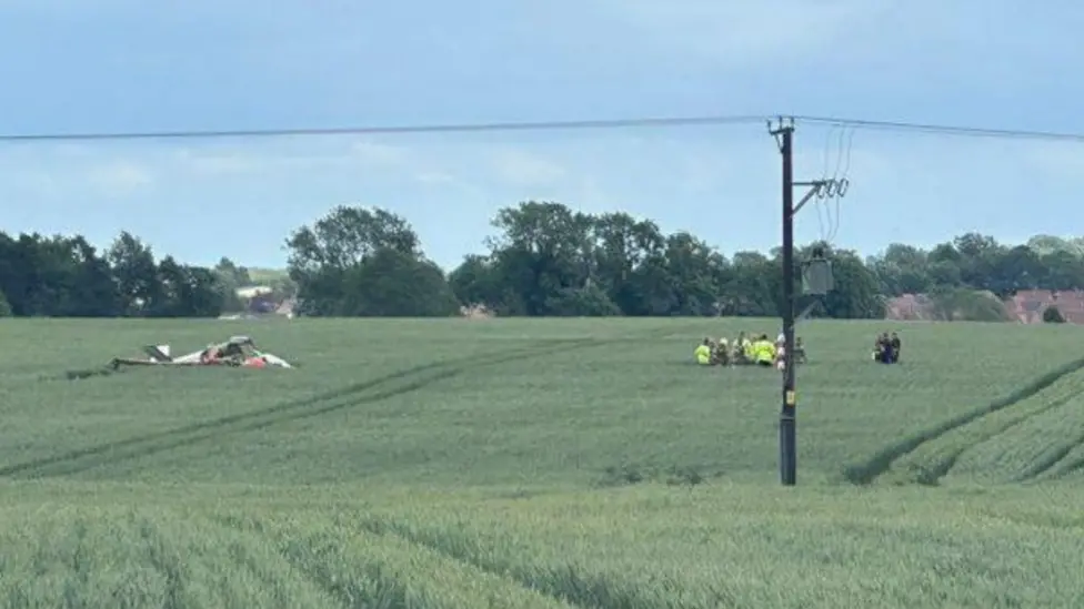 A small aircraft crashed on an open crop field, seen on the left, emergency response teams in high visibility jackets seen on the right. Trees and a blue, cloudy sky seen in the background.