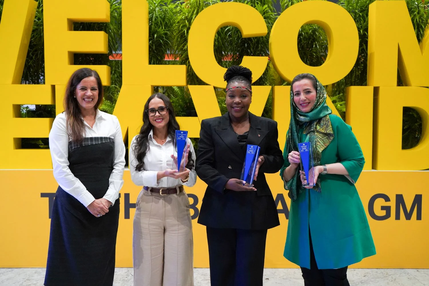 Group photo of four people standing side by side in front of a large, bright yellow backdrop with oversized letters spelling “WELCOME” partially visible behind them. Each person is dressed in formal or business attire and holds a tall, clear blue trophy with engraved text. One person on the left wears a black and white outfit, the next wears a white shirt with light trousers and glasses, the third wears a dark suit, and the fourth wears a green outfit with a matching headscarf. Lush green plants line the background above the sign, and the scene is brightly lit, suggesting a formal awards or recognition event.