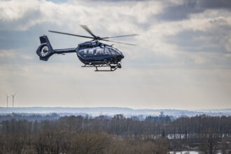 Helicopter flying above trees, under white clouds.