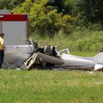 A small aircraft crash in a field, with a man standing next to it toward the left side. A fire truck is visible in the background.