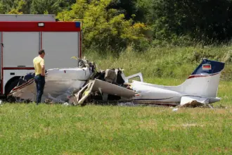A small aircraft crash in a field, with a man standing next to it toward the left side. A fire truck is visible in the background.