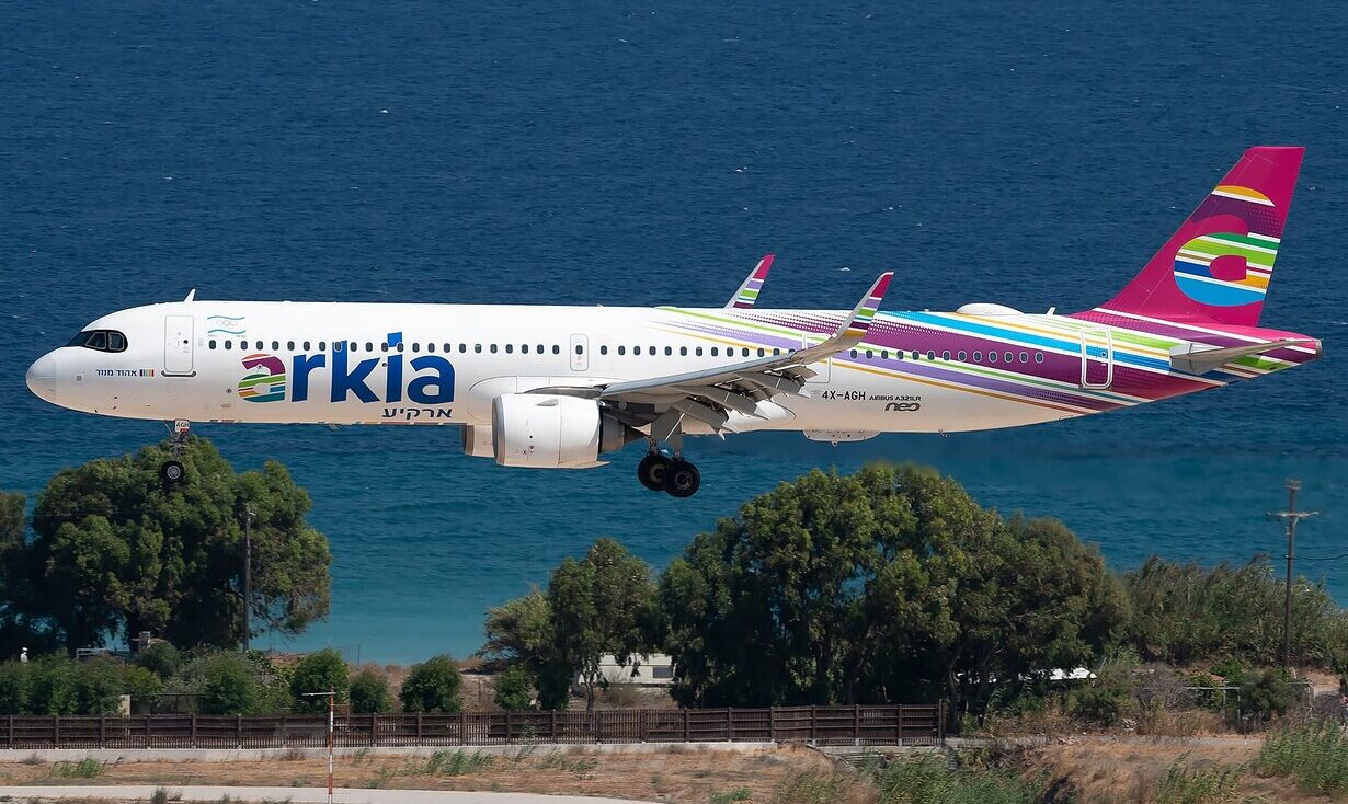 An Arkia Israeli Airlines Airbus A321neo flying in to land on a runway, its landing gear out. The ocean in the background, with trees visible below the aircraft.