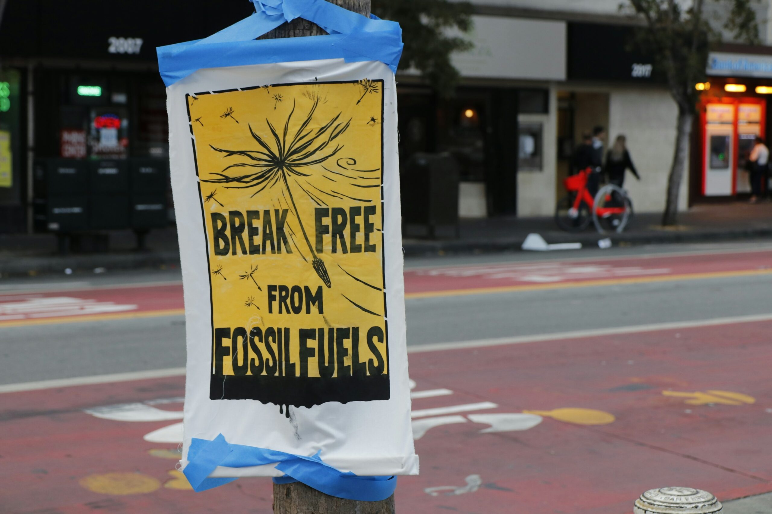 A sign is taped to a wooden post on the side of a road, reading 'BREAK FREE FROM FOSSIL FUELS'. The writing is in black, within a yellow rectangle with a black border, and the edges of the sign are white. Between the first two words is a drawing of a dandelion flying away in the same black ink. The sign is in the centre left of the image, and is the only thing in focus, with the background stylistically blurred. People can be vaguely seen walking on the opposite pavement, and a painted road can be seen immediately behind the sign.