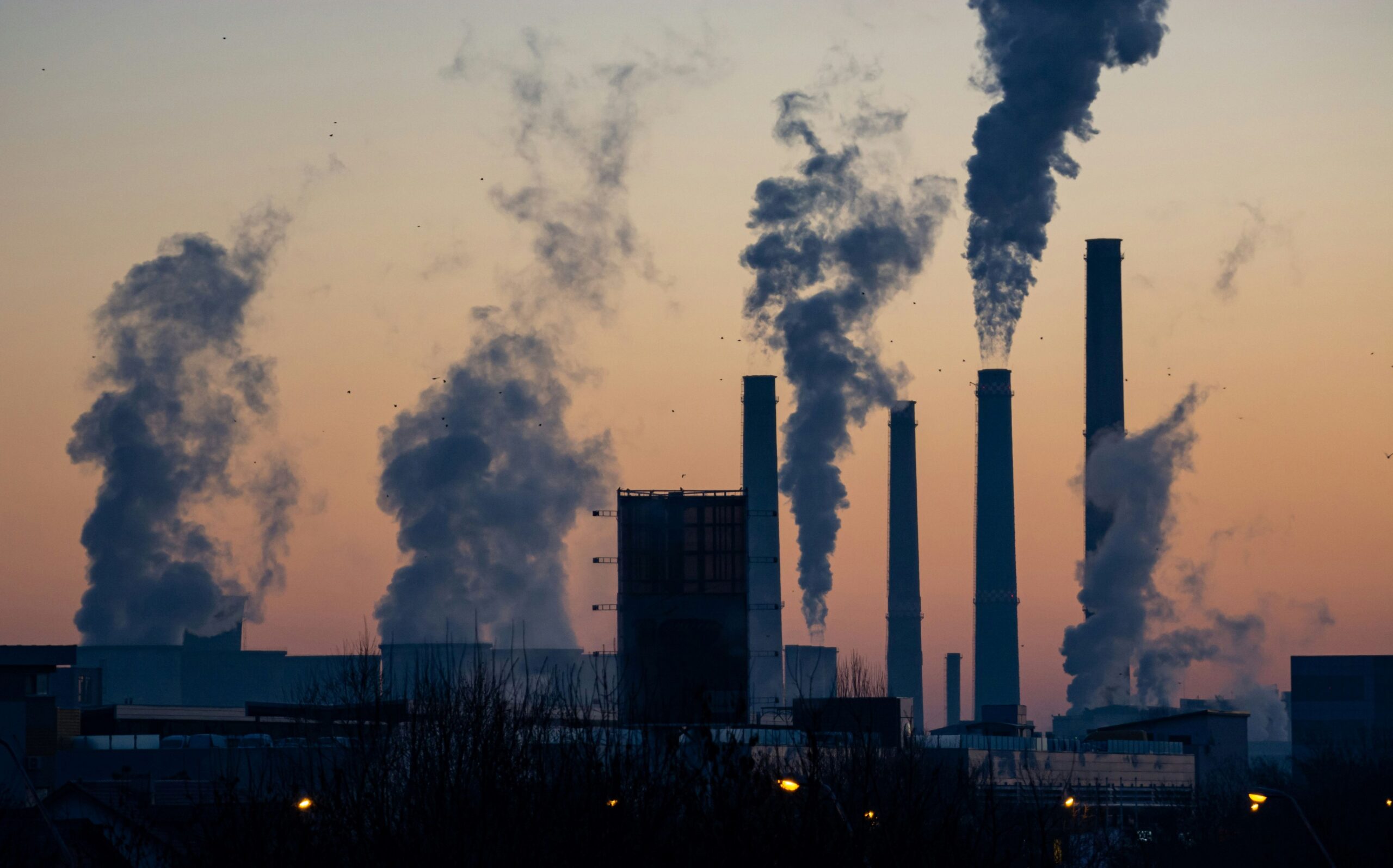 Fossil fuel production plant or factory from a distance at sunset. Large clouds of smoke can be seen rising from huge industrial sized chimneys. 