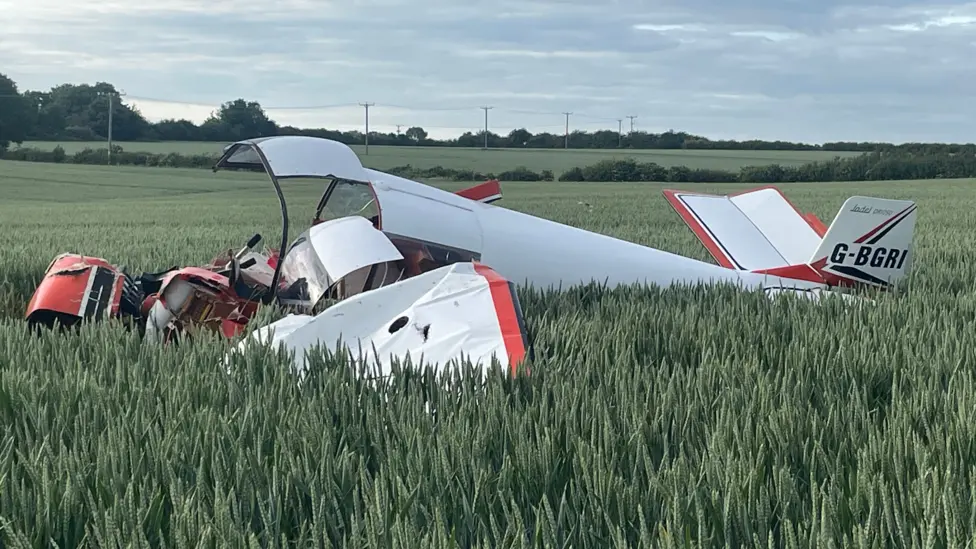 Small aircraft crash in a crop field, trees and cloudy sky in the background. The aircraft is upside down with the cockpit crushed. Red and white livery.