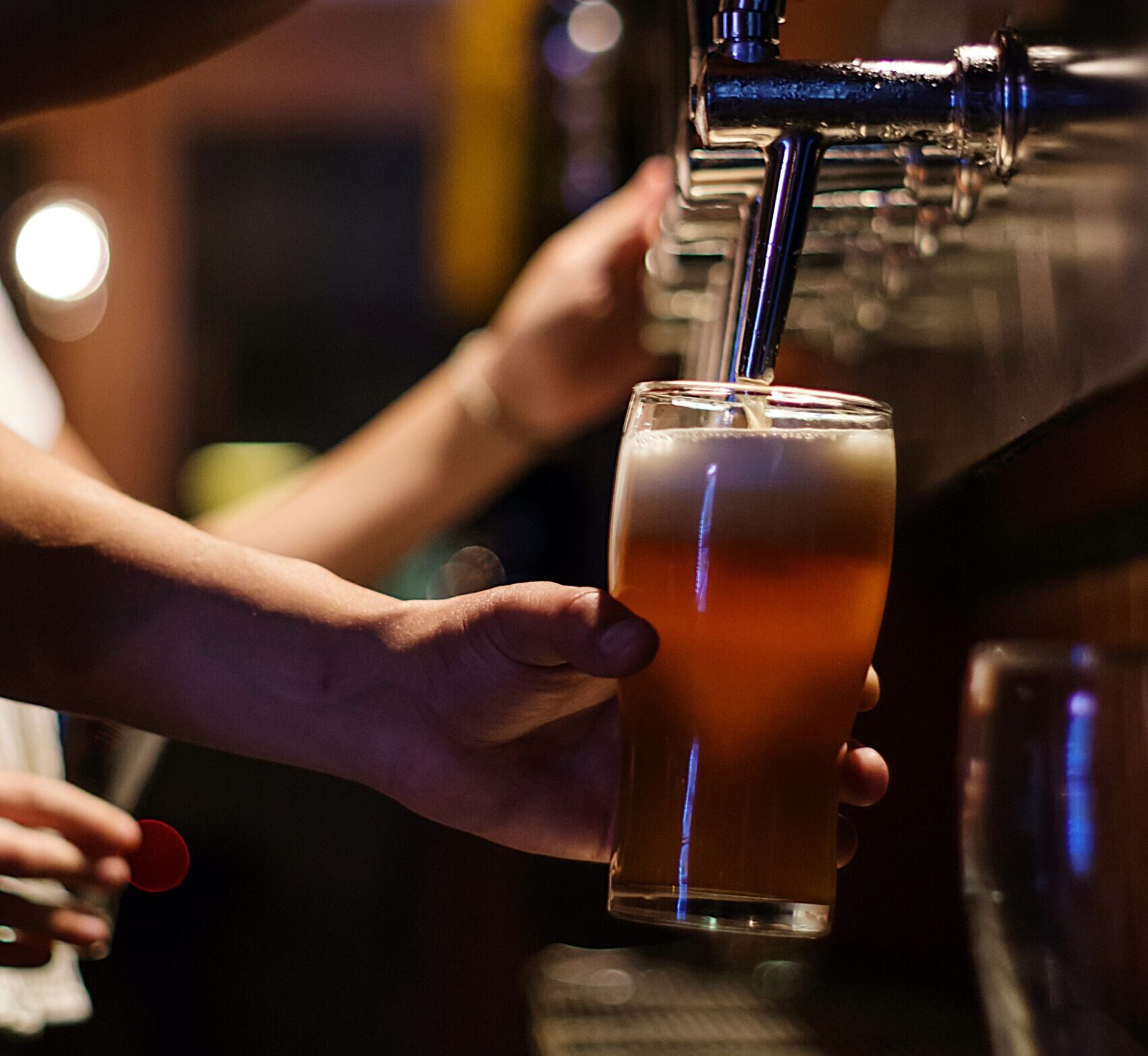 An image of a Caucasian hand holding a pint glass under a daught tap and pouring a pint of lager. The glass is a traditional shape, and is three quarters of the way full of beer. The background is stylistically blurred, but further daught taps and another Caucasian hand can be seen in the background.
