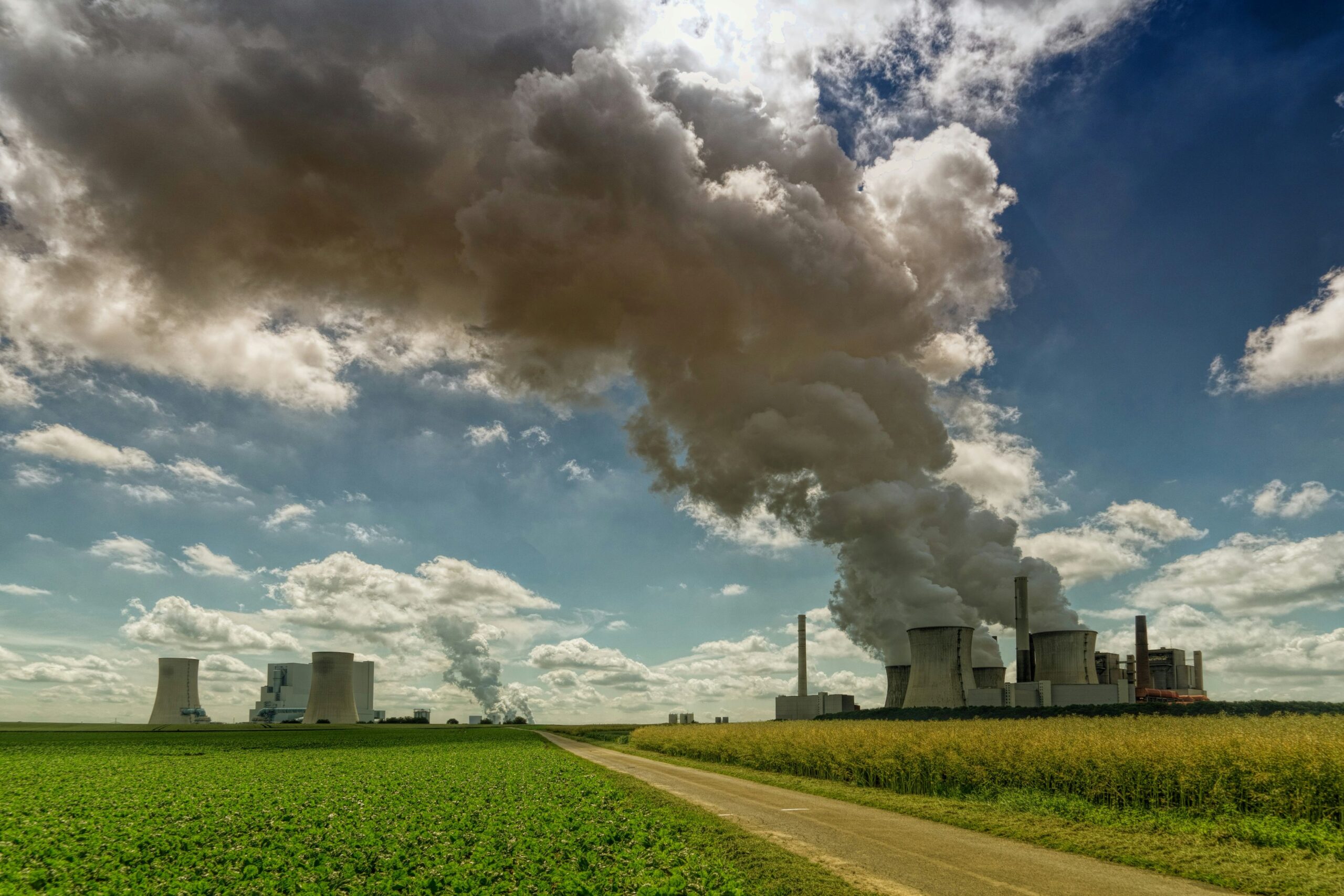 A large power plant can be seen in a field divided by a dirt road. Large plumes of smoke can be seen rising up from the huge white industrial style chimneys. The foreground is occupied mainly by green grass and the dirt road which travels from the right corner into the centre. The background is a large bue sky with clouds. 
