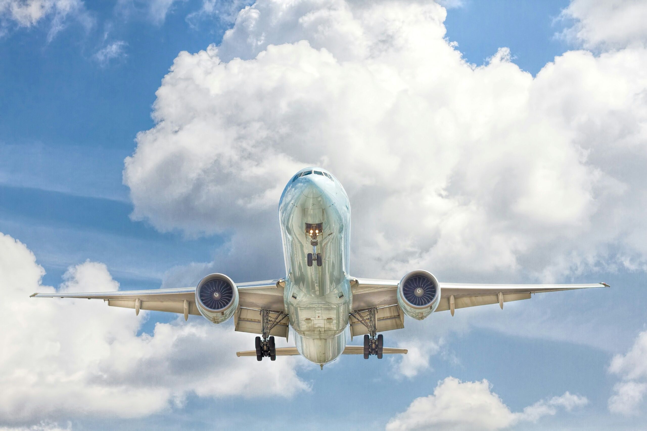 A large aircraft in flight in a blue sky with clouds from almost directly below. The jet, which is in the centre of the photo, is clean and so shiny it almost mirrors what is beneath it.
