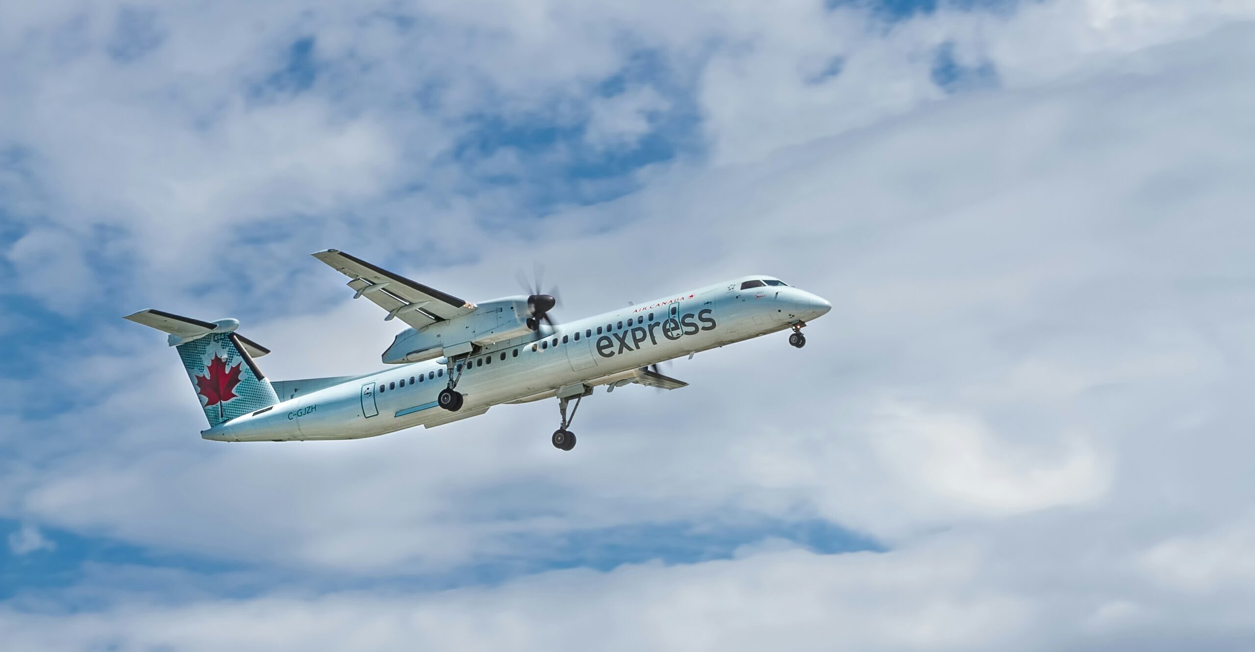A De Havilland Q400 is pictured in flight. The aircraft is centre left of the image against a background of cloudy blue sky. The aircraft is white with a red maple leaf logo of Air Canada on the tail, also with red and grey written branding on the side. The nose of the aircraft is facing slightly upwards and to the right, with the tail facing slightly downwards and to the left. 