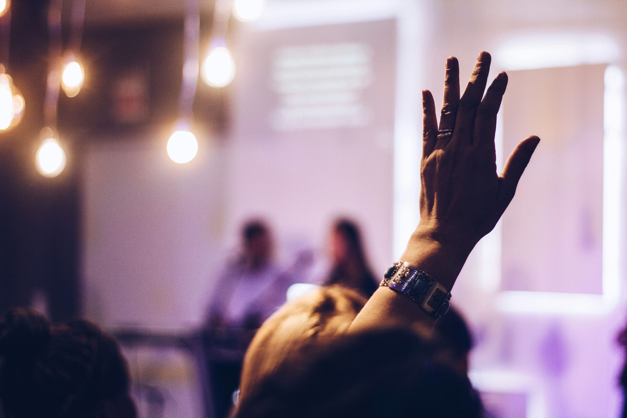 A raised hand is in focus in the centre right of the image, with everything else surrounding it stylistically blurred. Despite the blurring, it can be seen that the hand - which appears to be Caucasian and is adorned with a large silver bangle bracelet - is being raised by one person sat in a crowd attending some form of panellist talk and presentation. The lights and screen can be seen vaguelyin the background, and they give the image a warm purplish yellow undertone. 