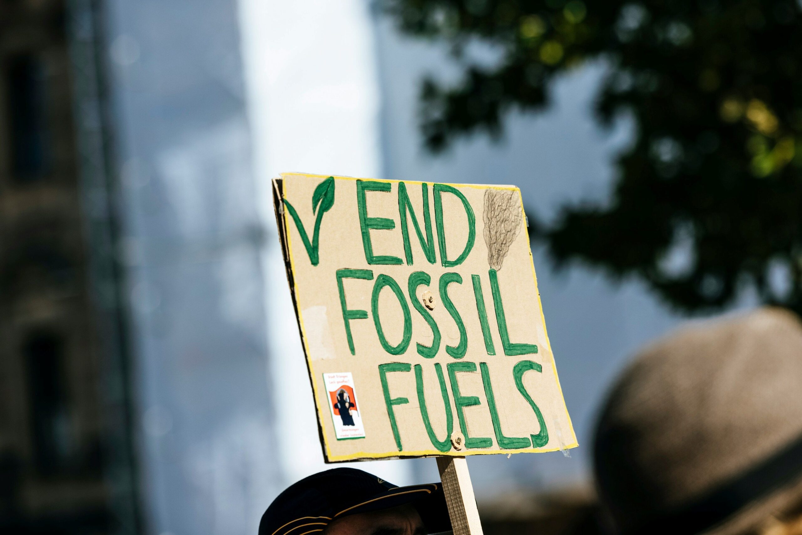 A cardboard sign in the air reading "End Fossil Fuels" in forest green paint. The background is stylistically blurred and appears to be held above people's heads 