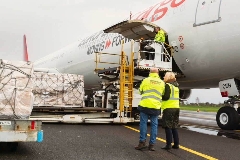 Ground crew in high-visibility jackets load cargo onto a European Cargo aircraft using a raised loading platform on an airport apron. Large wrapped freight pallets sit in the foreground as the aircraft’s cargo door is open under an overcast sky.