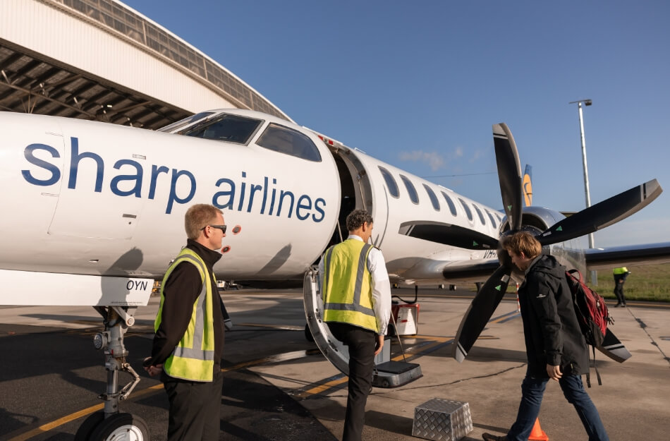 A Sharp Airlines aircraft parked on a runway. There are two workers with neon yellow vests and a passenger boarding the plane. The sky is blue.