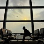 Silhouette of a man sitting at Kolkata Airport with a plane in the sky during sunset.