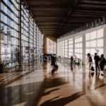 Passengers sit and stand in a busy airport departure lounge with carry-on luggage while checking electronic flight information boards displaying delays and gate information.
