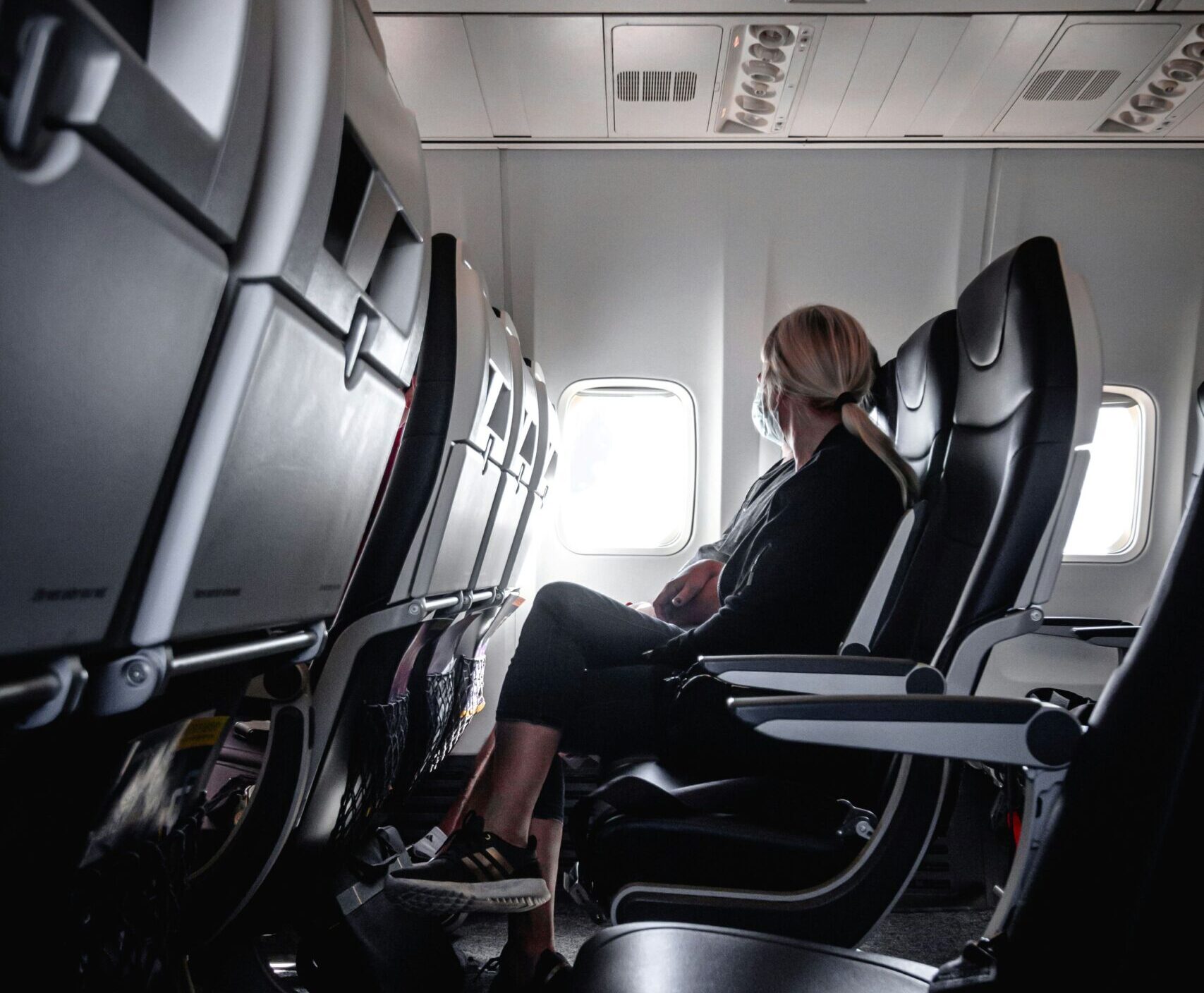 A female presenting person is sat in the middle seat of a row on an aircraft. The whole background is the greyish white inside of plane. The person is facing away from the camera and looking towards the window, with medium length blonde hair tied back in a low ponytail. The person is wearing charcoal greyish clothes and black shoes. The seats surrounding them also appear charcoal grey and light grey.