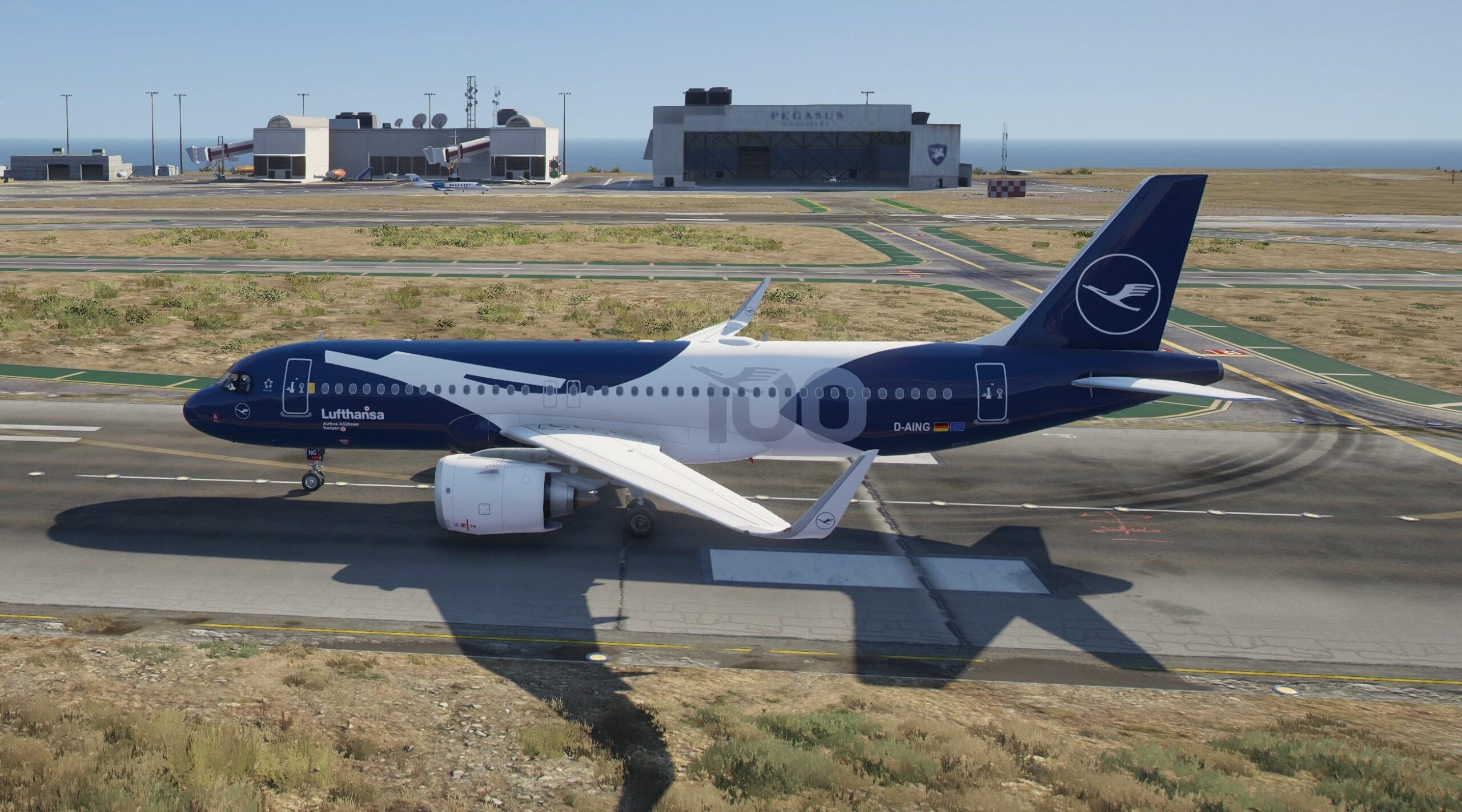 An Airbus A320neo aircraft with Lufthansa branding is stationed on the ground at a runway. It in the centre of the image, pictured directly from side on, the nose facing the left and the tail facing the right. is dark blue and white, and has special edition livery which includes '100' in light grey across side of back end. In the background operational buildings can be seen beyond the other runways and taxiway. The sky is blue and cloudless, and the ground is grey tarmac surrounded by yellowing grass.