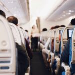 A stylistically blurred image of the inside of an aircraft taken from an aisle seat near the back. The aisle floor is dark blue and the back of the seats and the ceiling is white. the back of peoples heads can be seen in the image