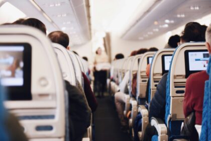 A stylistically blurred image of the inside of an aircraft taken from an aisle seat near the back. The aisle floor is dark blue and the back of the seats and the ceiling is white. the back of peoples heads can be seen in the image