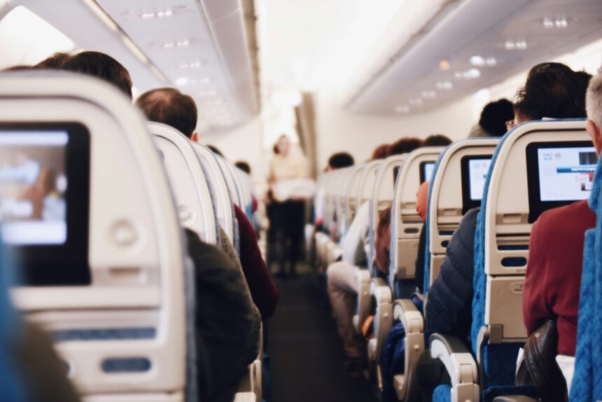 A stylistically blurred image of the inside of an aircraft taken from an aisle seat near the back. The aisle floor is dark blue and the back of the seats and the ceiling is white. the back of peoples heads can be seen in the image