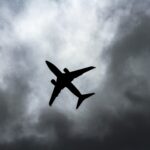 The shadowy silhouette of a aircraft in flight against a backdrop of stormy grey clouds. The picture is taken from a distance directly below. The aircraft is at an angle, with the tail facing towards the bottom right corner and the nose facing towards the top left corner.