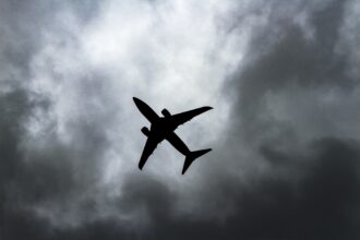The shadowy silhouette of a aircraft in flight against a backdrop of stormy grey clouds. The picture is taken from a distance directly below. The aircraft is at an angle, with the tail facing towards the bottom right corner and the nose facing towards the top left corner.