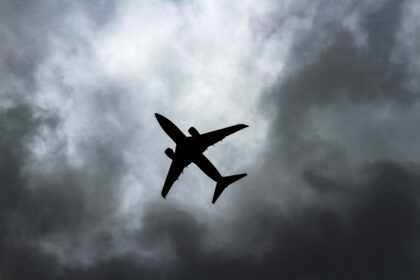 The shadowy silhouette of a aircraft in flight against a backdrop of stormy grey clouds. The picture is taken from a distance directly below. The aircraft is at an angle, with the tail facing towards the bottom right corner and the nose facing towards the top left corner.