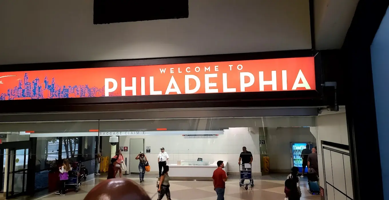 A "Welcome to Philadelphia" sign at PHL. Red background with white writing. Passengers below the sign walking around.