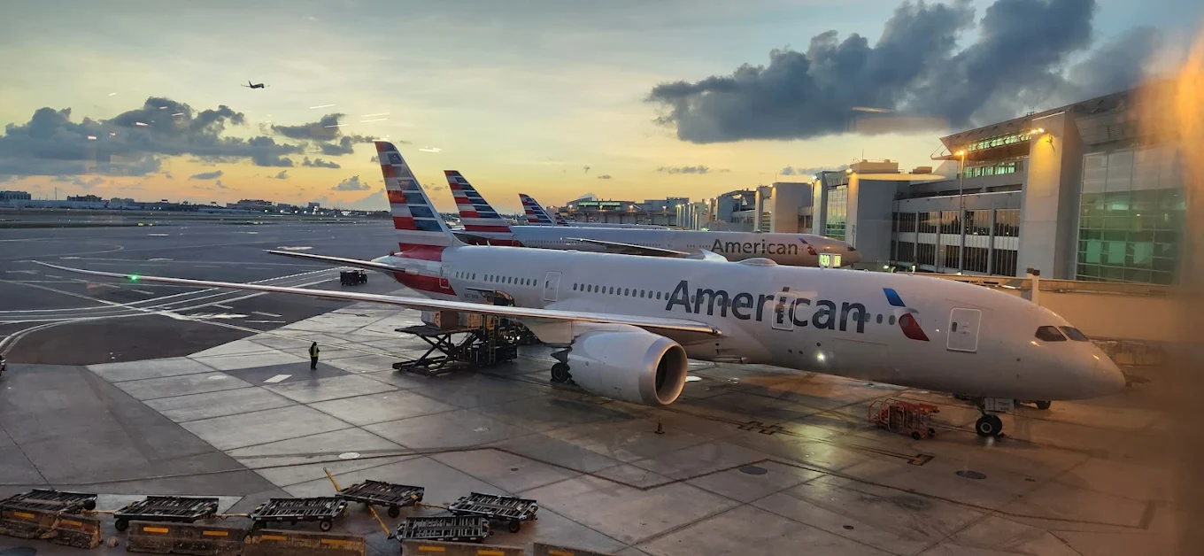 American Airlines aircraft parked at the apron in front of the airport building. Three aircraft visible on the ground while one is in the sky flying. The sun setting in the background with clouds.