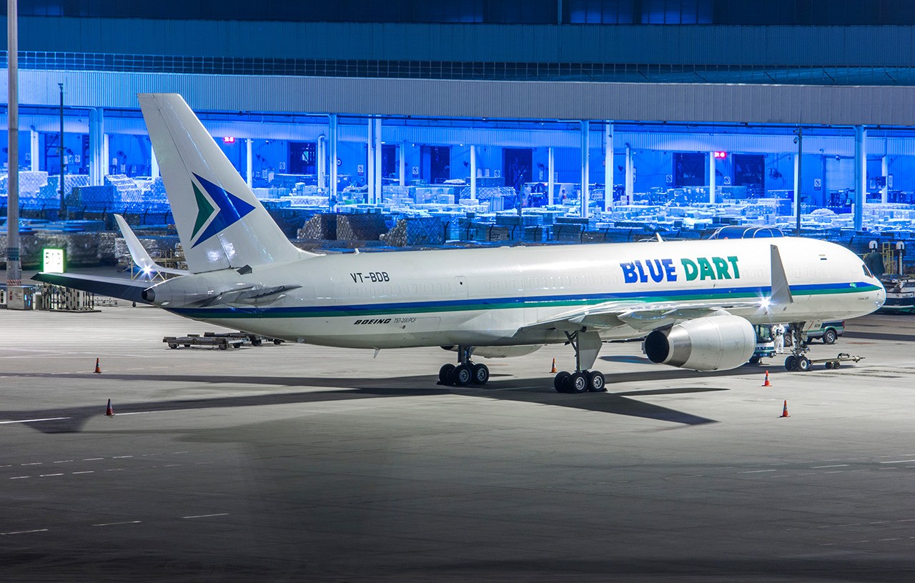 Blue Dart Aviation Boeing 757-200PCF freighter VT-BDB parked on the ramp at night with cargo containers visible in the background
