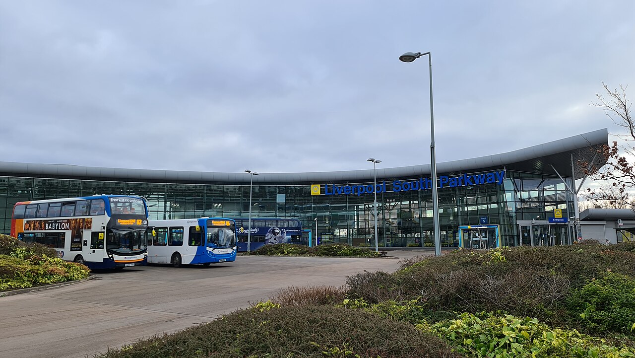 The outside of the Liverpool South Parkway railway station. The sky is cloudy with hints of blue. The building is mostly glass are there are two double decker buses parked next to the terminal.