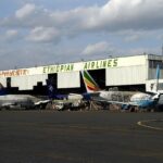 The picture depicts Bole International Airport in Addis Ababa, Ethiopia. There is a big hangar for aircraft shown with lettering saying 'Ethiopian Airlines' in a bright green font. Many Ethiopian aircraft are stationed in front of the hangar and waiting to be boarded.