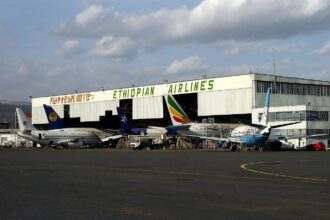 The picture depicts Bole International Airport in Addis Ababa, Ethiopia. There is a big hangar for aircraft shown with lettering saying 'Ethiopian Airlines' in a bright green font. Many Ethiopian aircraft are stationed in front of the hangar and waiting to be boarded.