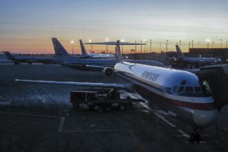 A series of American Airlines planes. The closest plane is silver with a black, red and white stripe all across the planes body. In the background is a sunrise with an orange to blue ombre.
