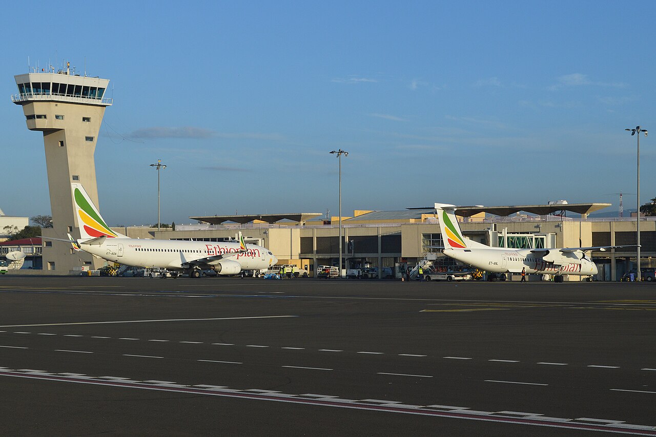 Two of Ethiopian Airlines fleet on stand at their home airport in Bole International Airport, Addis Ababa, Ethiopia. The background contains a sky that is bright blue, and a prominent feature is the airports control tower which stands to the side.