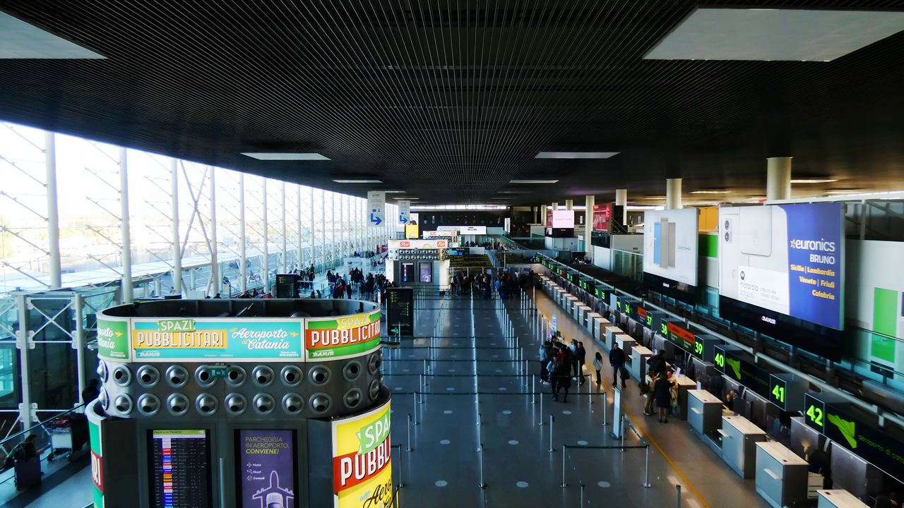 Departures and check in of Catania Airport. There are several check in desks, colourful billboards and one of the windows is covered in glass.