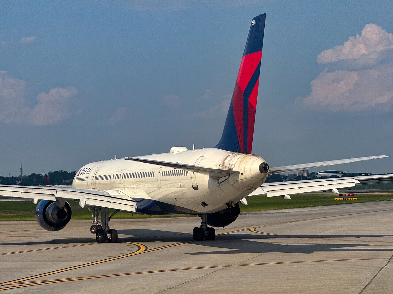 Photo depicts the side/rear end of a Delta airplane. It has a white body with blue and red accents on its rear wing. It's rear end has a distinctive point to it. In the background is a nice calm blue sky.
