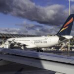 Image shows Plus Ultra Airbus A330 aircraft parked at Madrid Airport with dark clouds in a blue sky above, another aircraft wing in the foreground and airport vehicles nearby