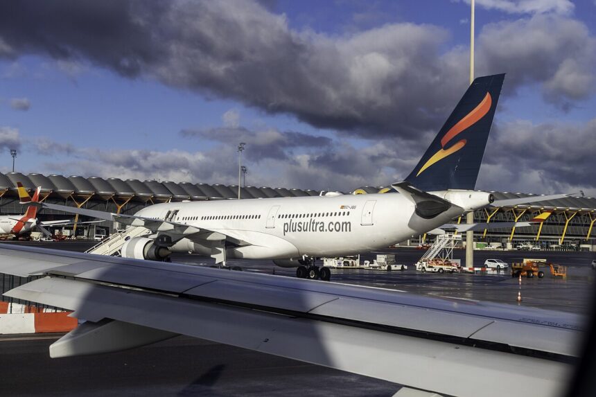 Image shows Plus Ultra Airbus A330 aircraft parked at Madrid Airport with dark clouds in a blue sky above, another aircraft wing in the foreground and airport vehicles nearby