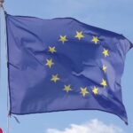 A picture of the dark blue with yellow stars European Union flag fluttering in the wind, with a bright light blue sky in the background.