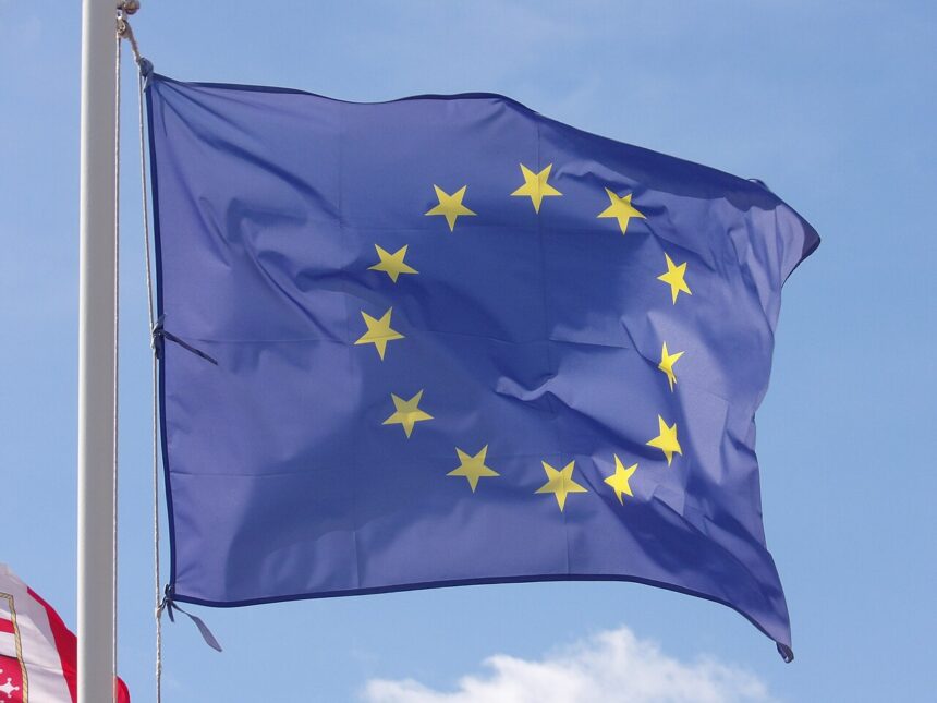 A picture of the dark blue with yellow stars European Union flag fluttering in the wind, with a bright light blue sky in the background.