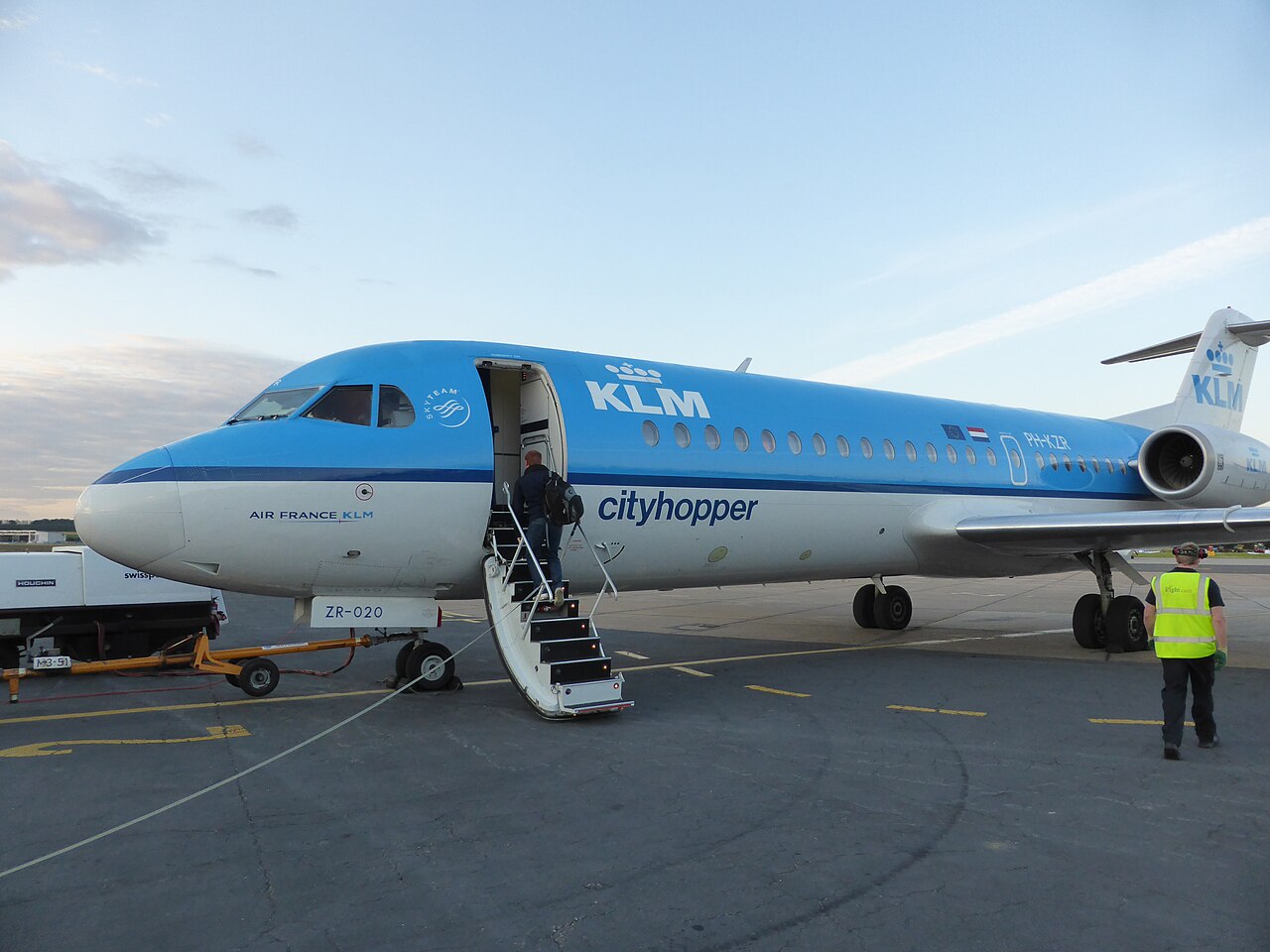A picture of a light blue and white KLM Cityhopper with its door wide open and a man boarding at Humberside airport. There is a airport worker in a high visibility vest casually walking to the back of the plane. The sky is bright blue with some clouds.