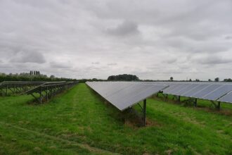 The photo depicts a grey cloudy day with solar panels lined up faced towards the sky. The grass is a lush green and trees are gathered in the distance.