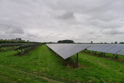 The photo depicts a grey cloudy day with solar panels lined up faced towards the sky. The grass is a lush green and trees are gathered in the distance.