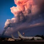 Mount Etna while erupting. In front of the volcano, there are US Navy aircraft on the ground. The sky is blue with a massive cloud of smoke.