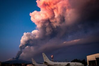 Mount Etna while erupting. In front of the volcano, there are US Navy aircraft on the ground. The sky is blue with a massive cloud of smoke.