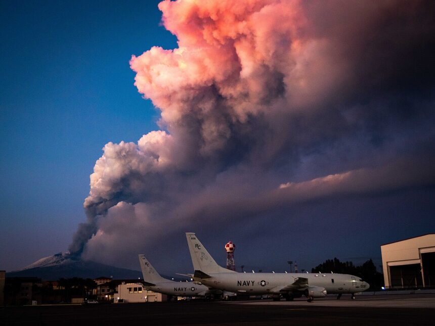 Mount Etna while erupting. In front of the volcano, there are US Navy aircraft on the ground. The sky is blue with a massive cloud of smoke.