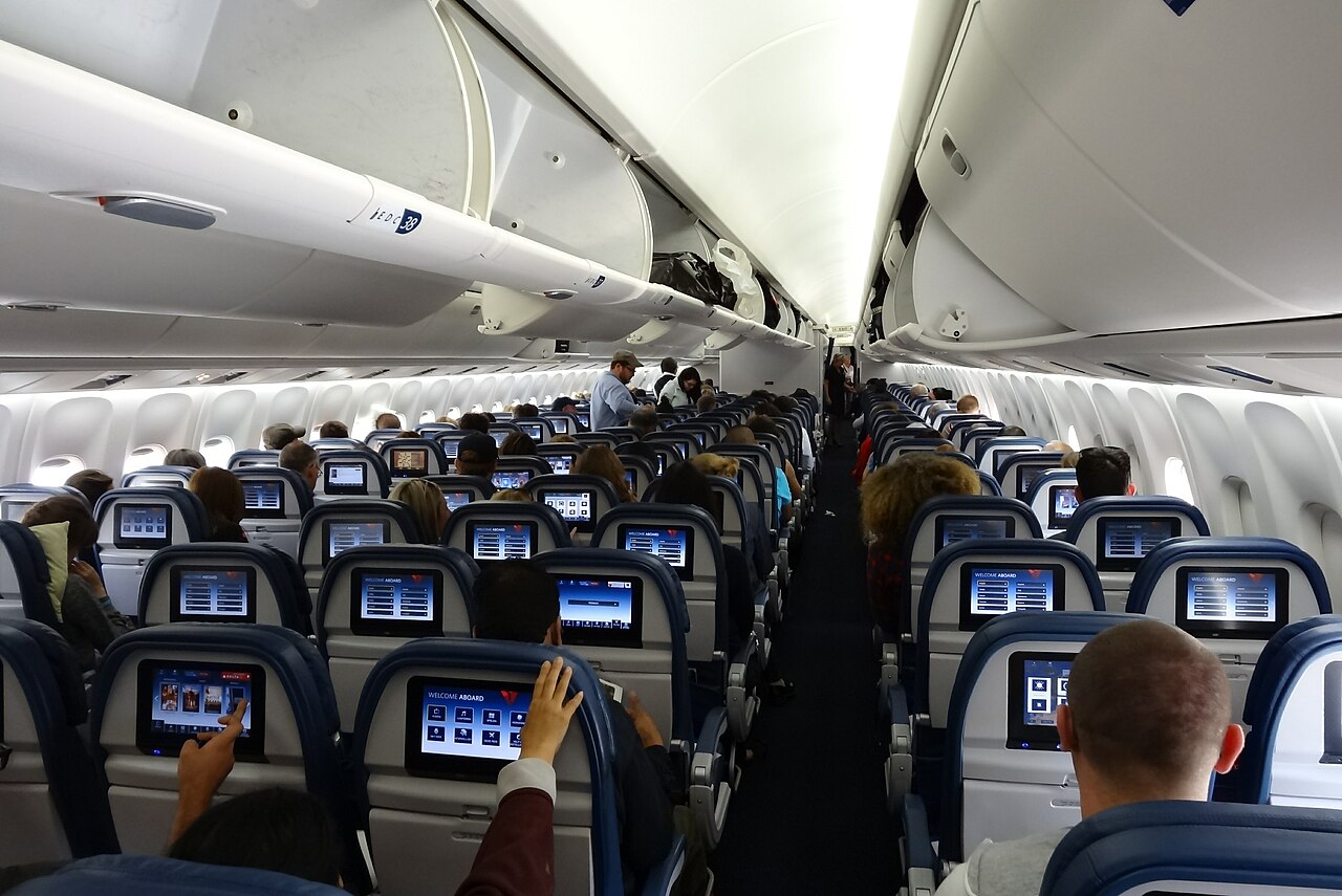 The photo depicts the inside of a Delta Air Line Boeing 767 looking down the plane. The heads of people sitting down are engaged in the mini screens, on the backs of the chairs in front of them. The cabin is encased in a calm white light.